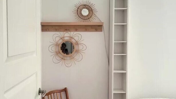Una stanza con una sedia di legno, un letto bianco, uno scaffale bianco con oggetti decorativi e un armadio bianco.