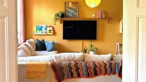 Una camera accogliente con un letto, una TV a schermo piatto montata al muro, una sveglia sopra il letto e una pianta sul davanzale.