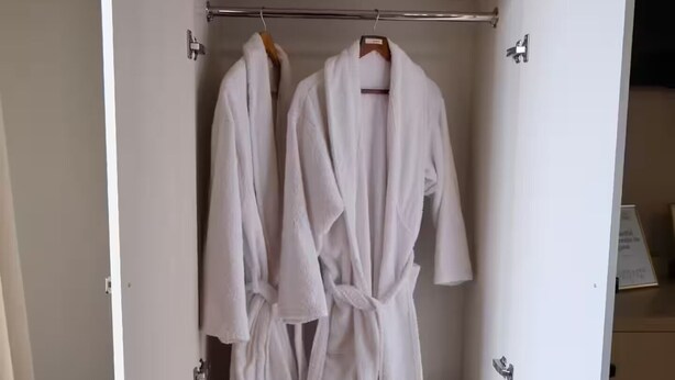 Um guarda-roupa com duas batas brancas penduradas em uma barra, uma prateleira com uma toalha branca dobrada e outra prateleira com um par de chinelos brancos.