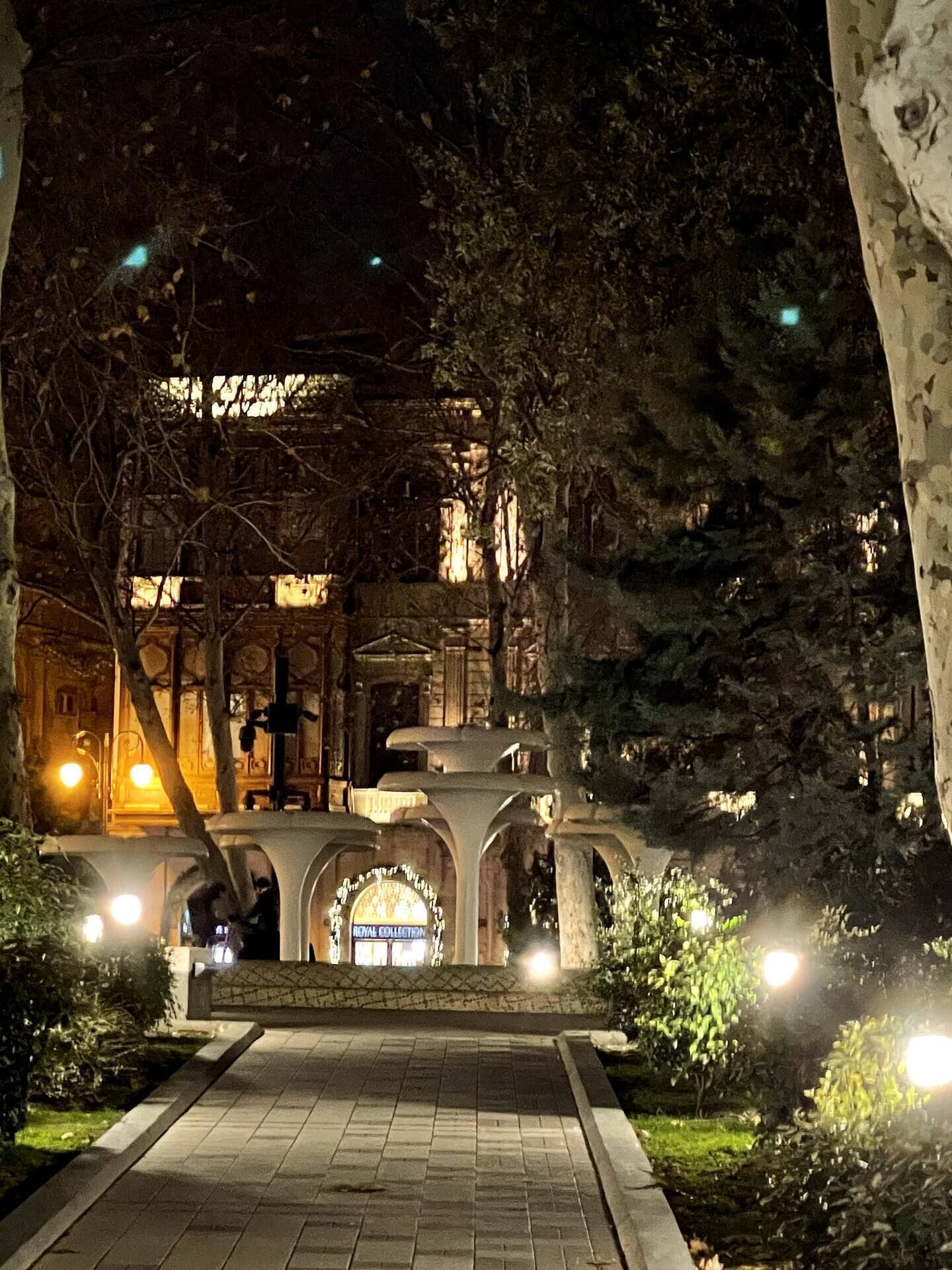 A nighttime view of a building with a central arched entrance, surrounded by trees and illuminated by street lamps.