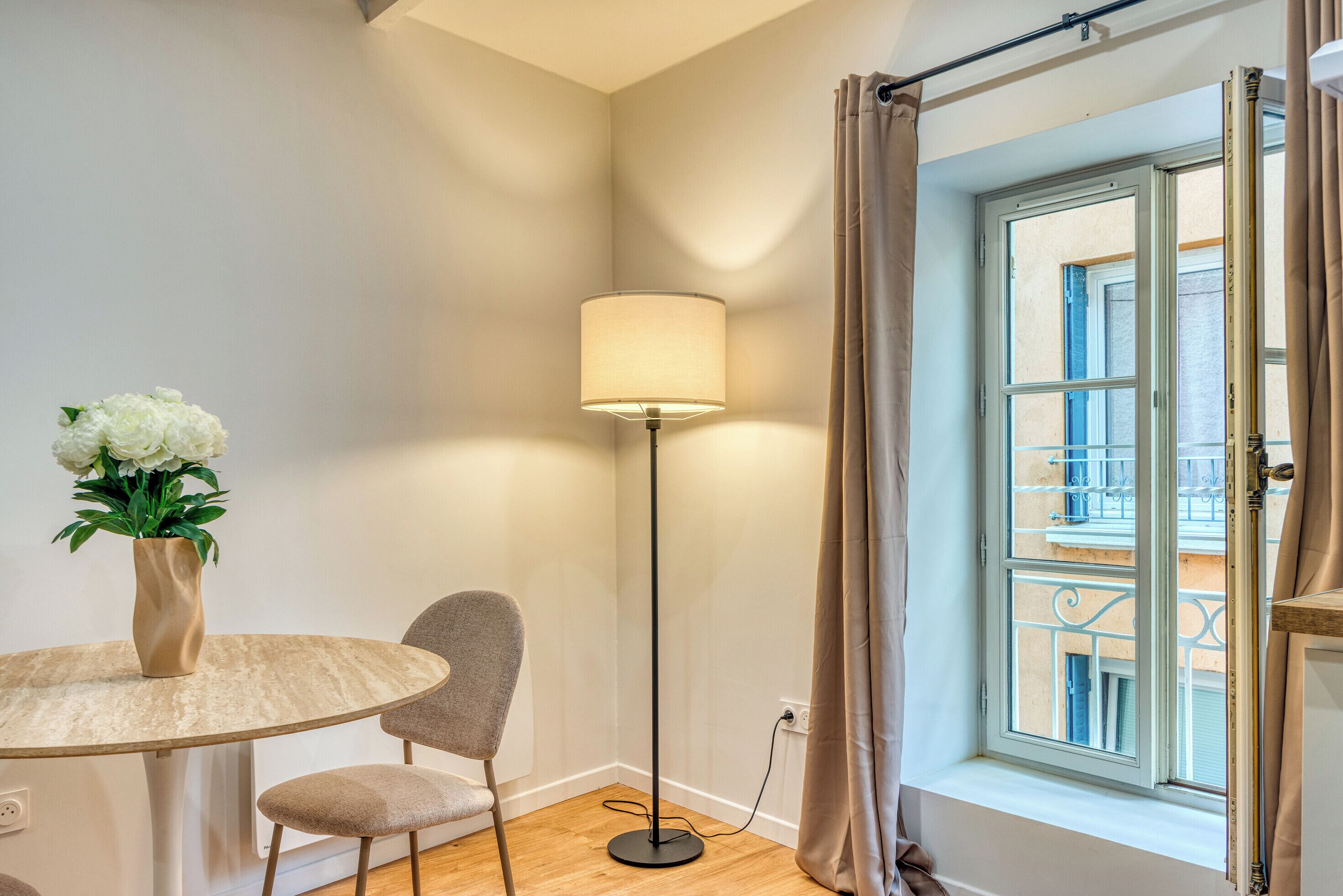 A corner room with a round table, a floor lamp, a potted plant, and a window with curtains.