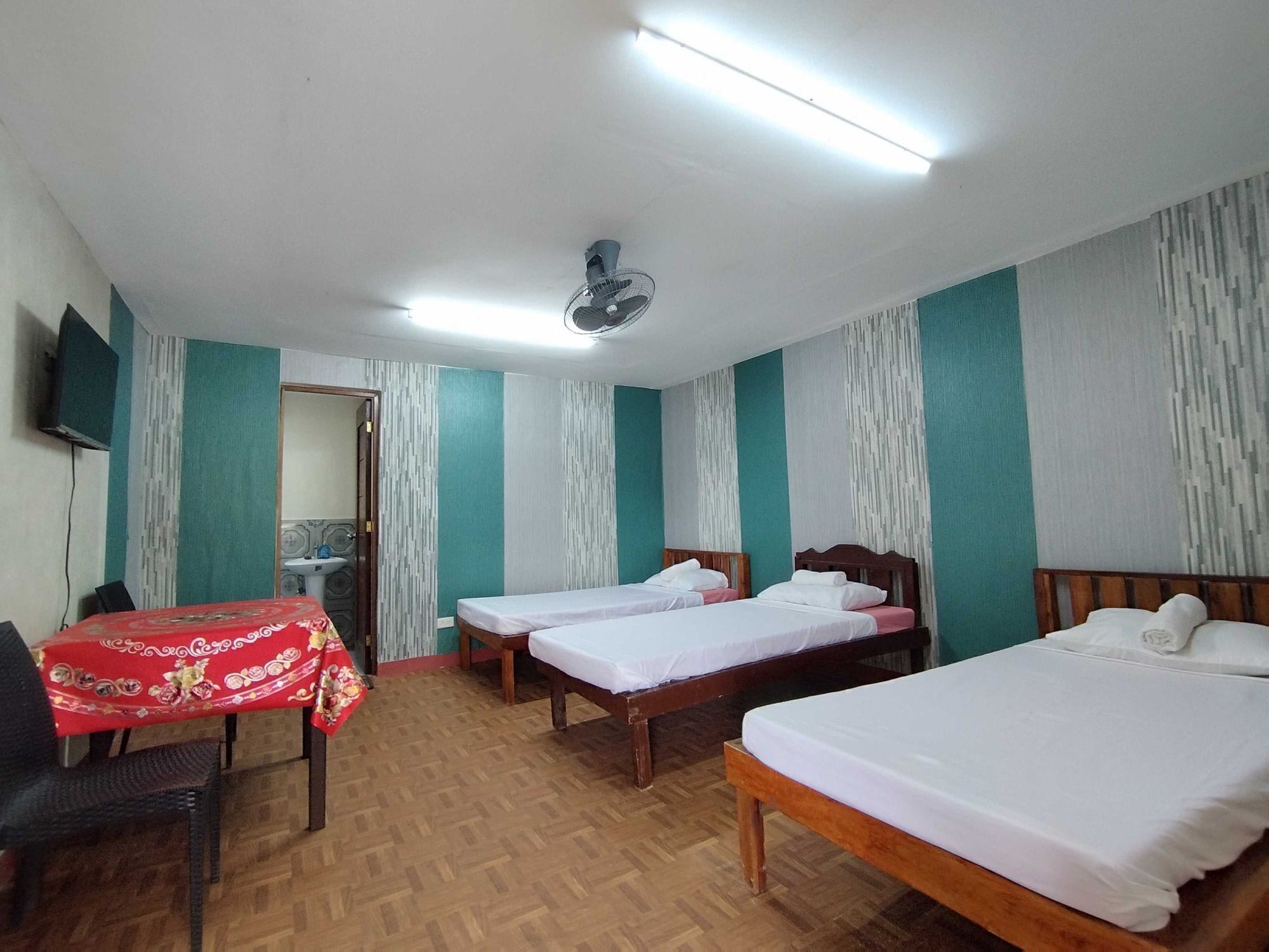 A hotel room with two single beds, a wooden floor, a red table with a patterned tablecloth, a ceiling fan, and a television mounted on the wall.