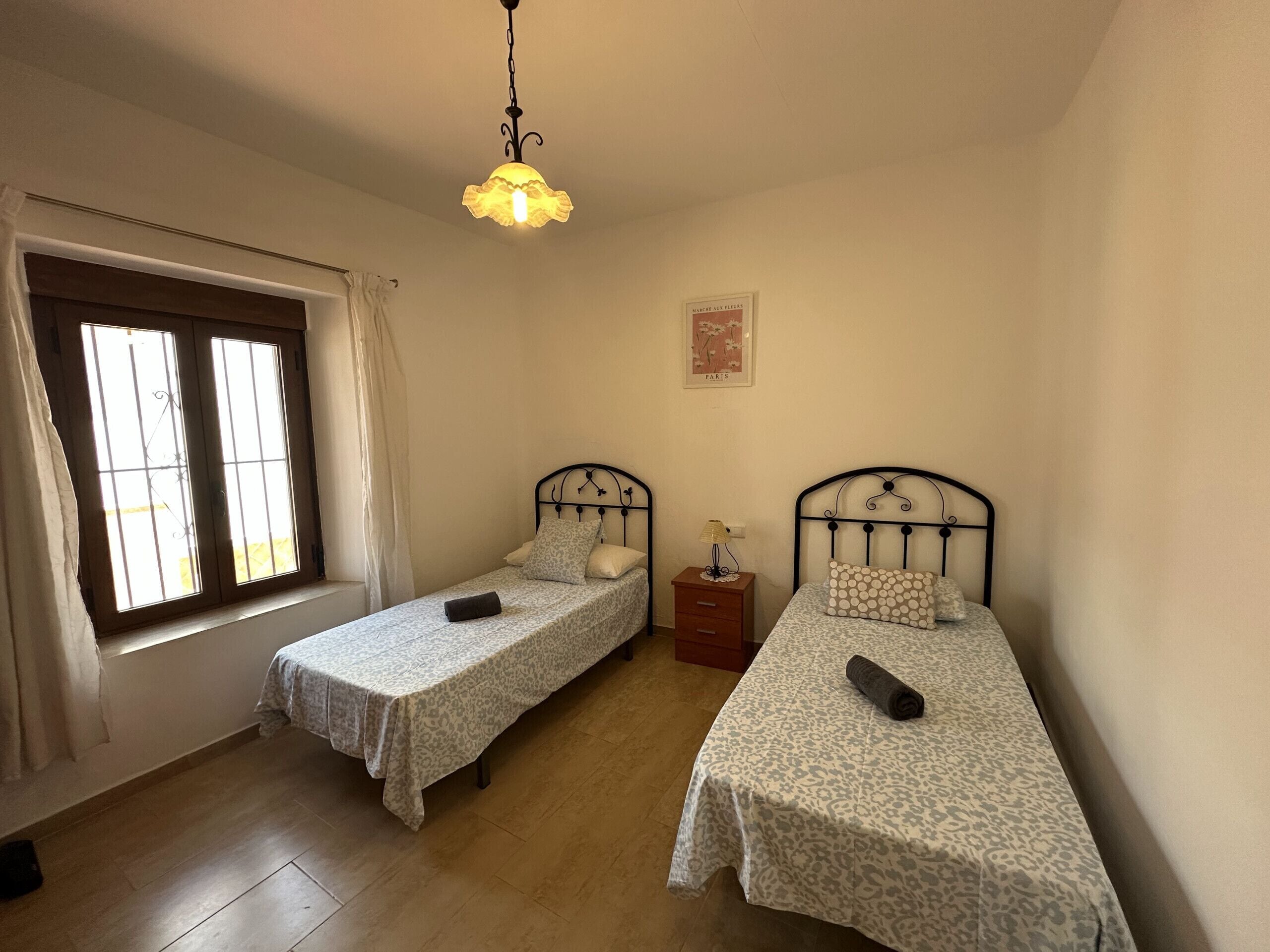 A room with two single beds, a window with curtains, a hanging light fixture, and a small bedside table with a lamp.
