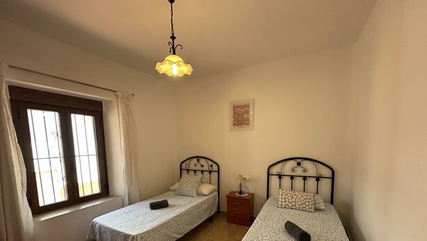 Una habitación con dos camas individuales, una ventana con cortinas, una lámpara colgante y una mesita de noche con lámpara.