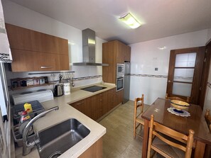 Een kleine keuken met houten keukenkastjes, een roestvrijstalen spoelbak, een inbouwoven en een eethoek met een houten tafel en stoelen.