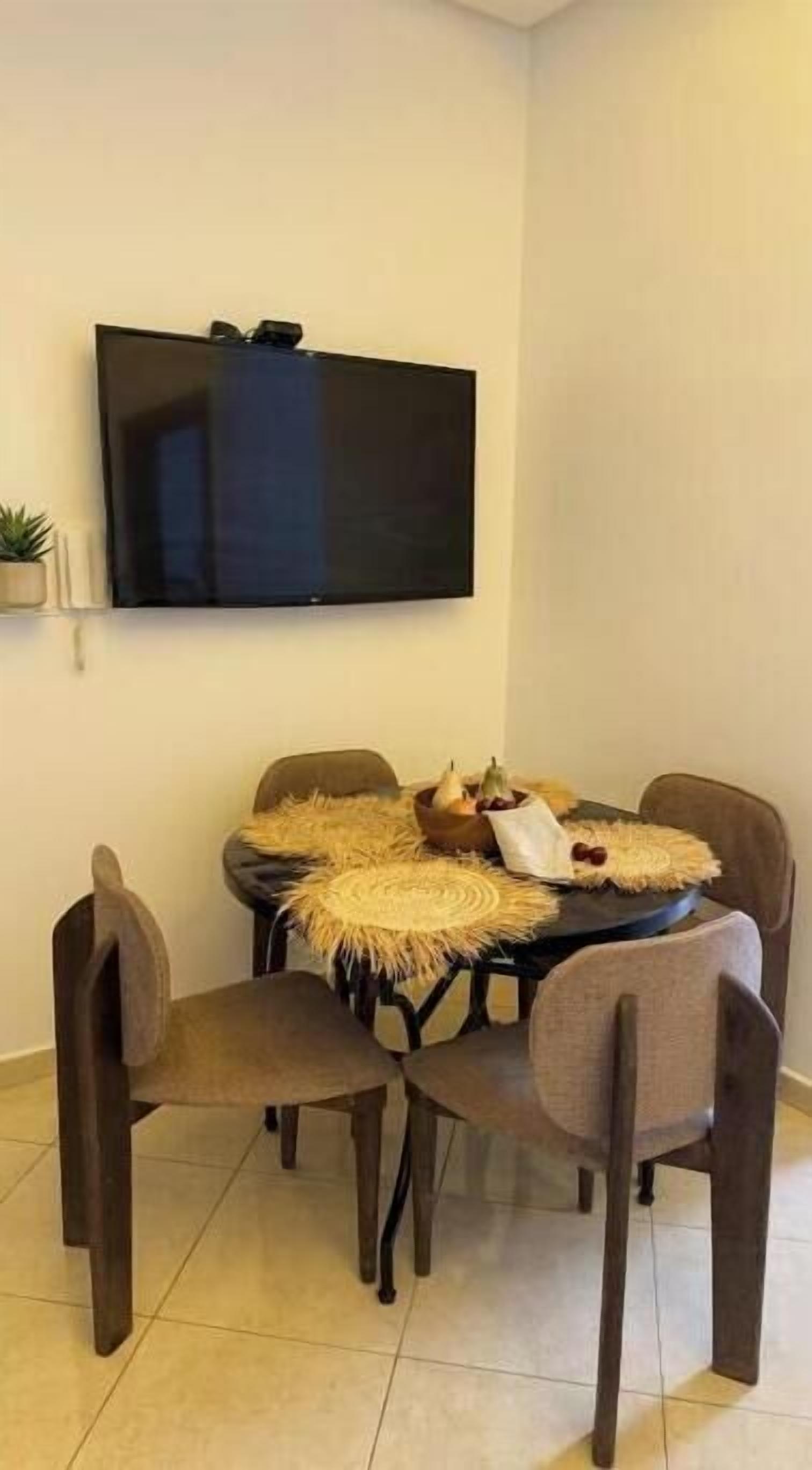 Un comedor con una televisión montada en la pared, una mesa redonda con cuatro sillas y una pequeña planta en maceta en la pared.