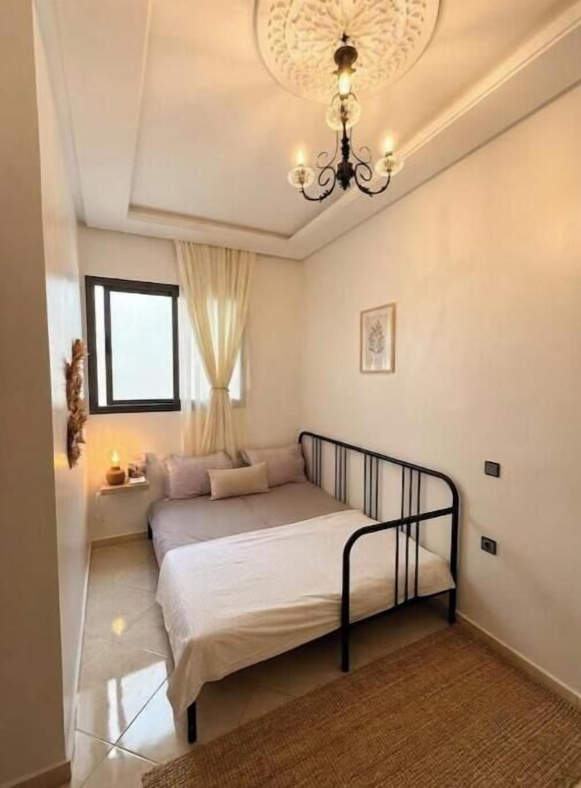 Um quarto com uma cama, um candelabro, uma janela com cortinas e uma mesa de cabeceira.