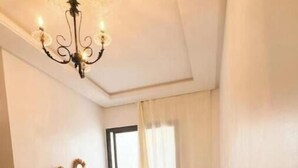 A bedroom with a bed, a bedside table, a chandelier, a window with curtains, and wall decorations.
