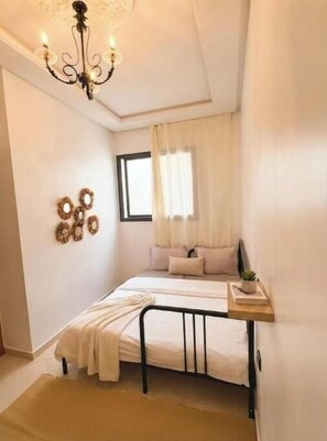 A bedroom with a bed, a bedside table, a chandelier, a window with curtains, and wall decorations.
