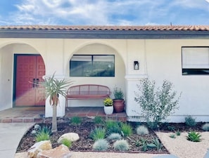 Una casa con puerta roja, un banco y un jardín con diversas plantas.