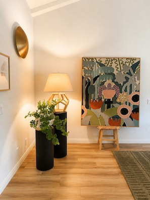 Una habitación con piso de madera, una lámpara de pie negra, un cuadro enmarco, un cuadro abstracto grande y dos plantas en macetas.