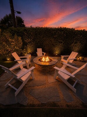 A fire pit surrounded by white Adirondack chairs in a landscaped outdoor area. - Stylish Designer Home Near Downtown & Bike Path (Ojai)