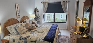 Ein Schlafzimmer mit einem Bett, einem Nachttisch, einer Lampe, einem Fenster mit Vorhängen und einem an der Wand befestigten Regal.