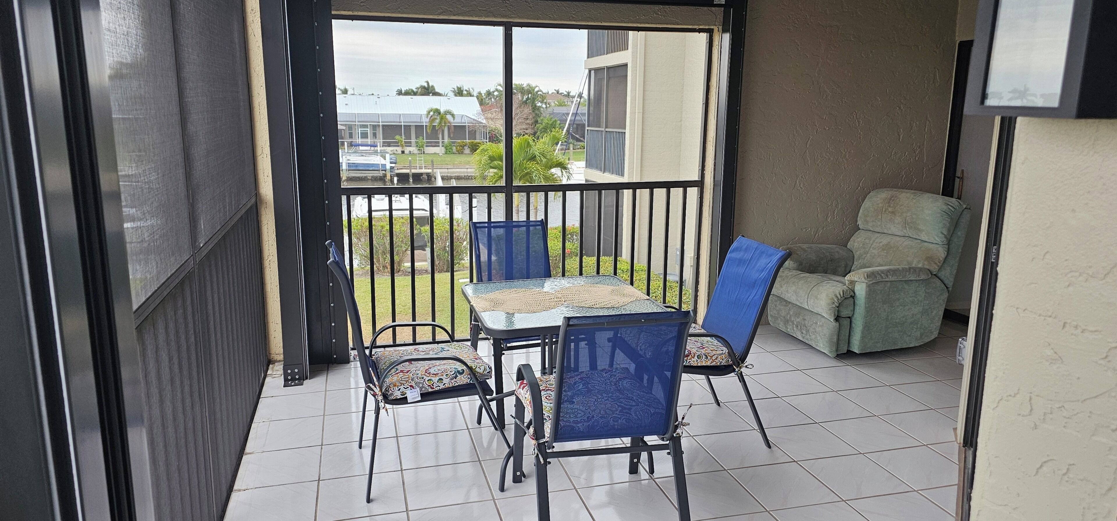 A covered patio area with a table and chairs, a cushioned chair, and a view of a building and greenery.