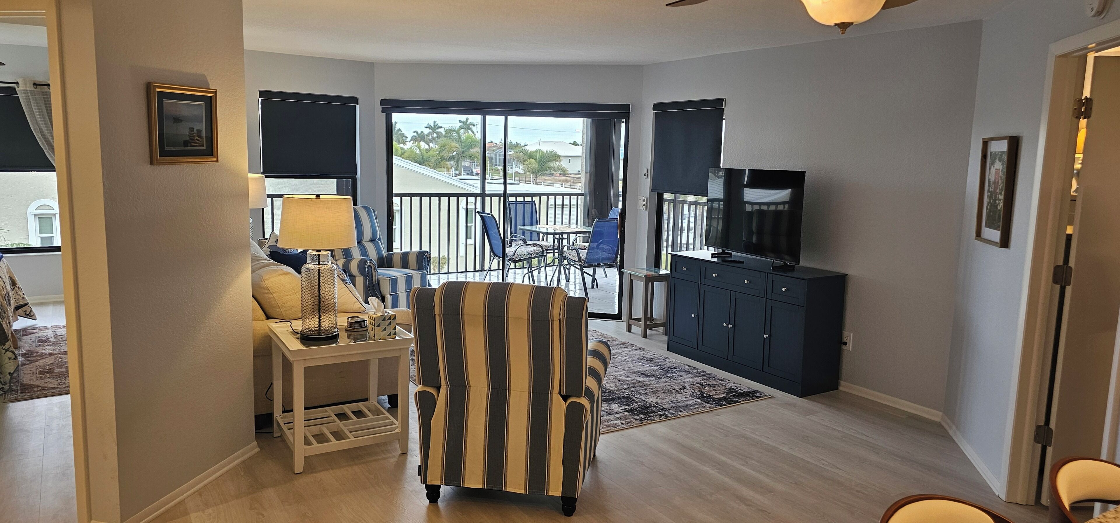 A living room with a striped armchair, a small table with a lamp, a TV on a cabinet, and a balcony with chairs.