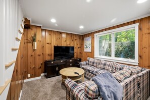 Ein gemütliches Wohnzimmer mit Holzwandverkleidung, einem Flachbildfernseher, einem karierten Sofa und einem großen Fenster.