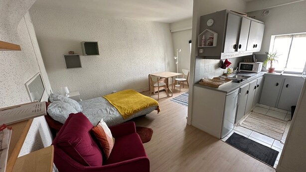 Ein gemütlicher, kompakter Wohnraum mit Bett, einem roten Sessel, einem kleinen Esstisch und einer Küchenzeile.