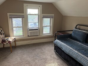 Een kamer met een schuine plafond, een bed met een blauw-witte streep dekbedovertrek, een stoel met een bloemenmotief en twee ramen met airconditionings.'