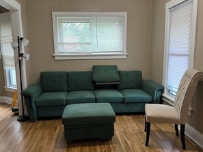 A living room with a green sofa, an ottoman, a chair, and a floor lamp. There is a window with blinds and a white wall. - Fantastic 3-bedroom house with AC in charming Benson Omaha (Omaha)