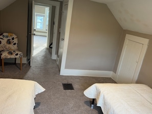 Een kamer met een schuine kap, een deur naar een gang, een gestreept stoel en twee bedden met witte dekens.