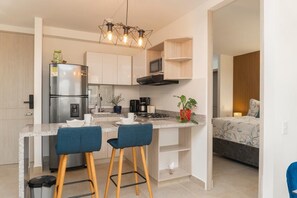 Eine moderne Küche mit integriertem Kühlschrank, Mikrowelle und Herd, ein Essbereich mit zwei Barhocker und ein Schlafzimmer mit Bett und Nachttisch.