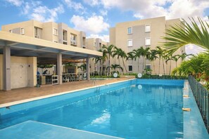 Una piscina con zona techada, palmeras y un edificio con balcones.