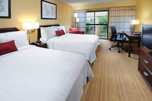 Ein Hotelzimmer mit zwei Betten, einem Schreibtisch, einem Sessel, einem Fernseher und einem Fenster mit Blick auf Grünflächen.
