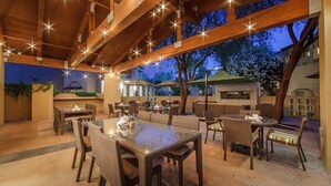 Een terras met houten balken en lichtslingers, omringd door tafels en stoelen.
