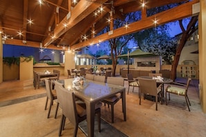 Un área de comedor al aire libre con vigas de madera y luces colgantes, rodeada de mesas y sillas.