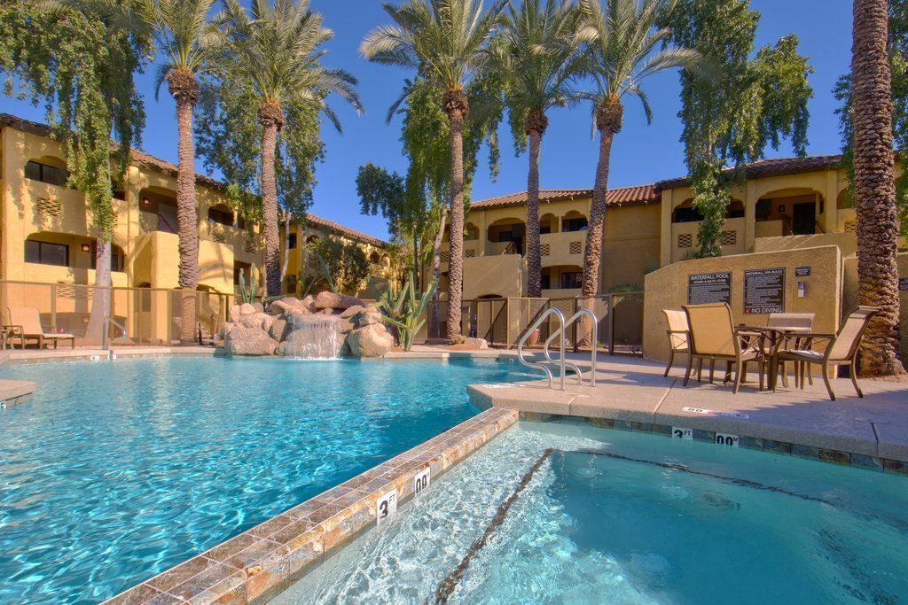 A swimming pool with clear blue water, surrounded by palm trees and a building with a tiled roof.