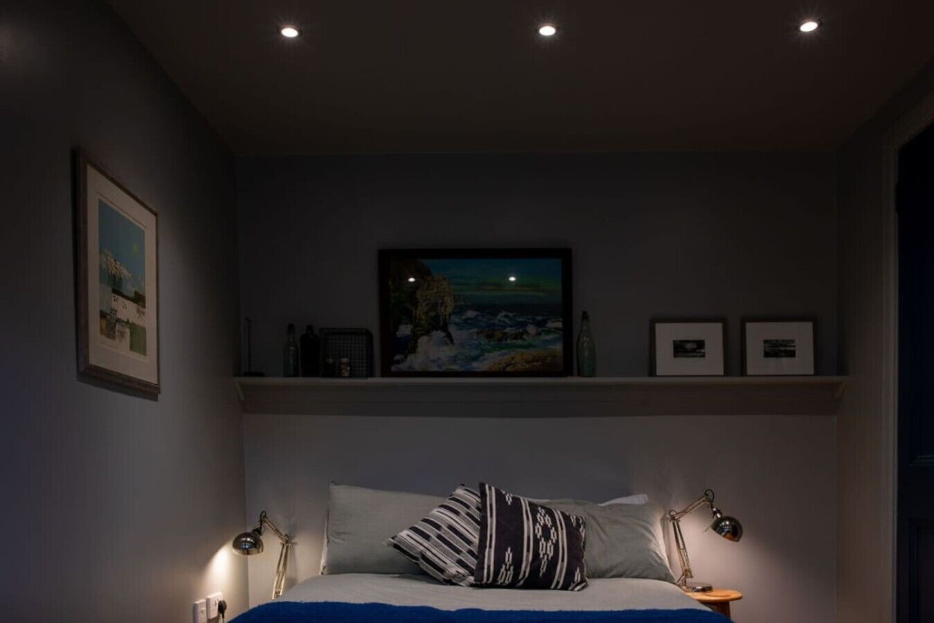 A bedroom with a bed, two bedside lamps, a nightstand, a shelf with framed pictures, and a painting above the bed.