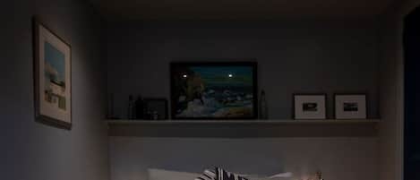 A bedroom with a bed, two bedside lamps, a nightstand, a shelf with framed pictures, and a painting above the bed.