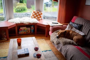 A cozy living room with a dog resting on a sofa, a coffee table with a book and glasses, and a window with a view of the outside.
