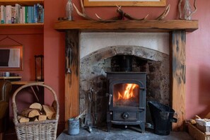 Una chimenea con fuego, una estantería, un sillón de mimbre y una cesta de leña.