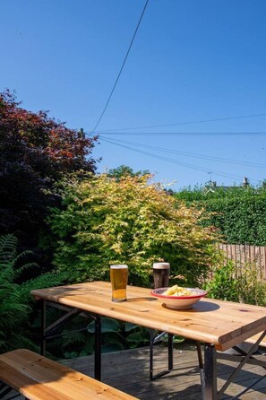 Une table en bois avec deux verres de bière et un bol de nourriture, disposée à l’extérieur, avec de la verdure et un ciel dégagé en arrière-plan.