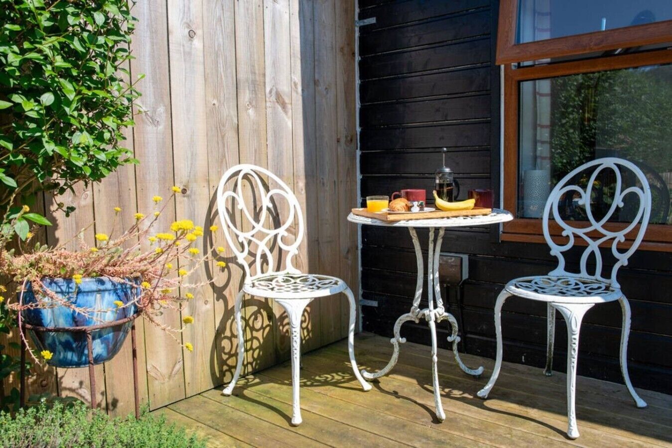 A wooden deck with two white metal chairs and a small round table, a hanging blue planter with yellow flowers, and a window with a view of greenery.