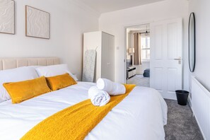 A hotel room with a bed, a white headboard, a yellow throw blanket, and a black trash bin. - Luxe 4-bedroom house perfect for groups | great transport links! (London)