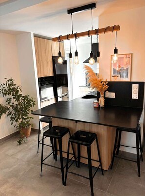 A modern kitchen with a central island, black bar stools, hanging pendant lights, and a potted plant. - APPARTEMENT 4 COUCHAGES PROCHE DU CENTRE (SIX FOURS LES PLAGES)