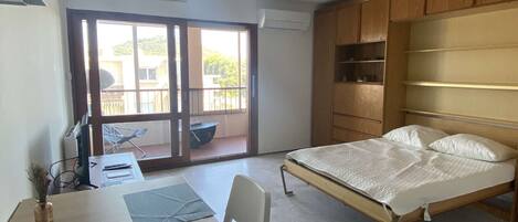 Un dormitorio con cama, escritorio, silla, armario y balcón.