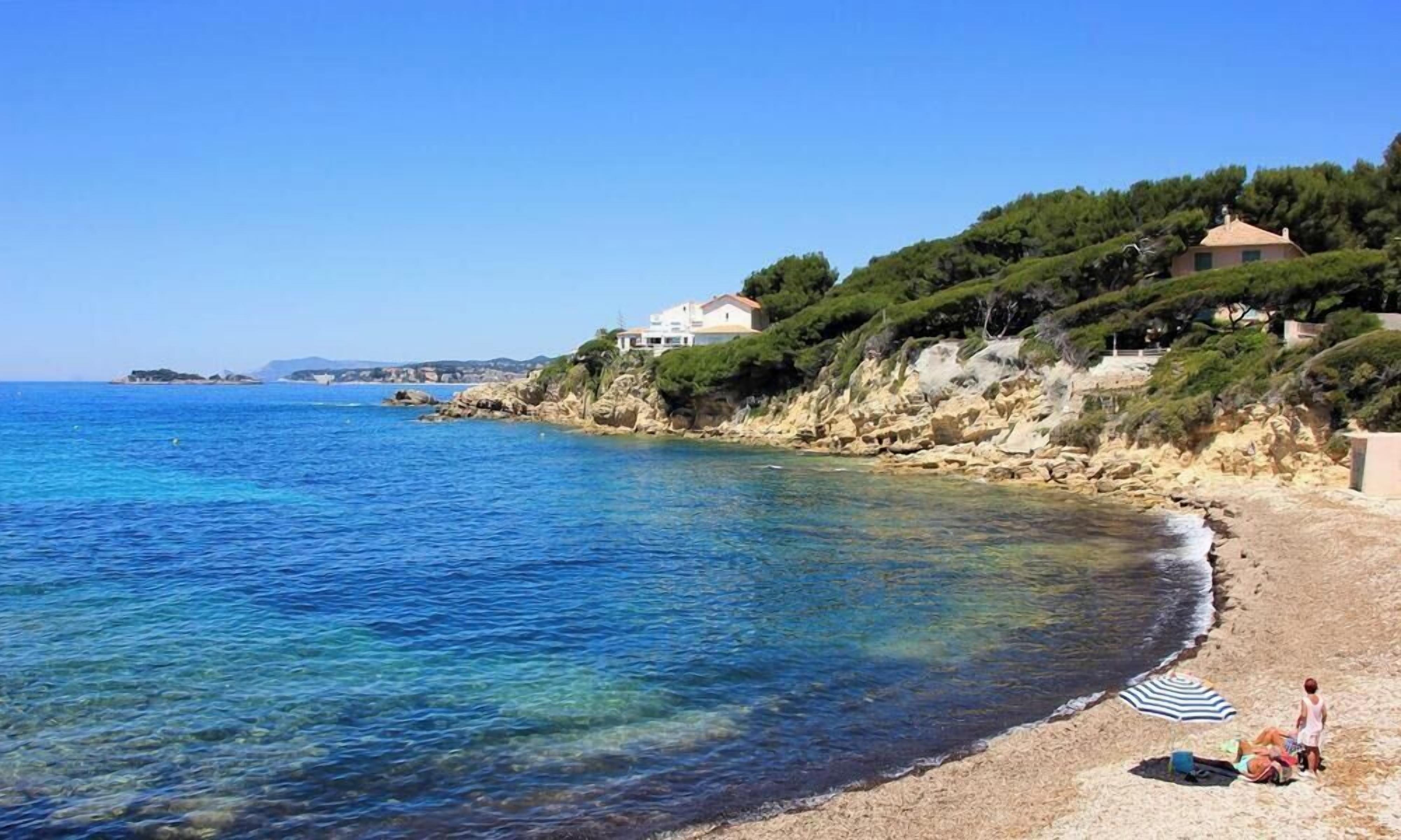 A coastal beach with clear water, a sandy shore, and houses on a hillside.