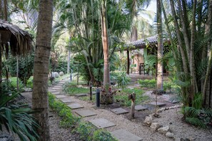 Um jardim tropical com um caminho de pedra, palmeiras e um edifício rústico com telhado de palha.
