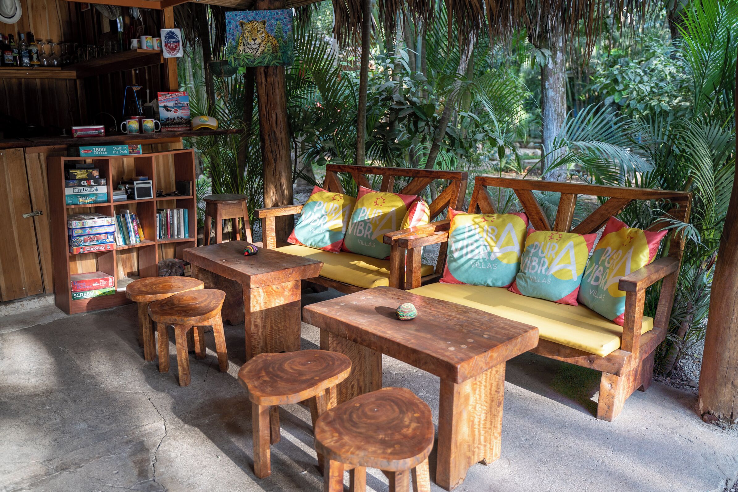 A wooden outdoor seating area with a sofa, tables, and chairs, surrounded by lush greenery.