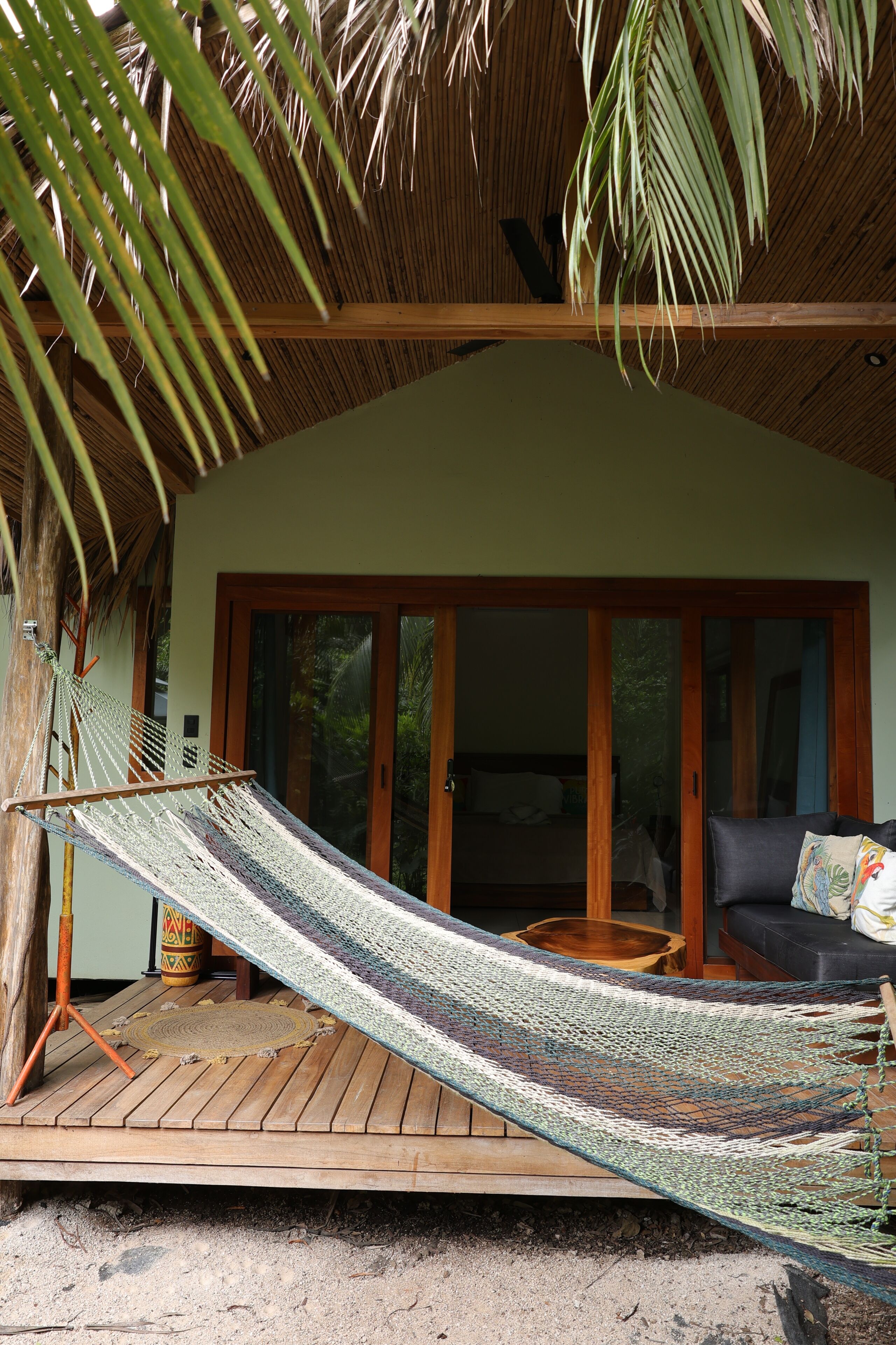 Una hamaca bajo un techo de madera, una terraza de madera y un ventanal corredizo que da a una habitación con cama.
