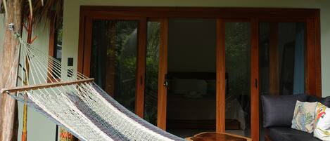 Una hamaca bajo un techo de madera, una terraza de madera y un ventanal corredizo que da a una habitación con cama.