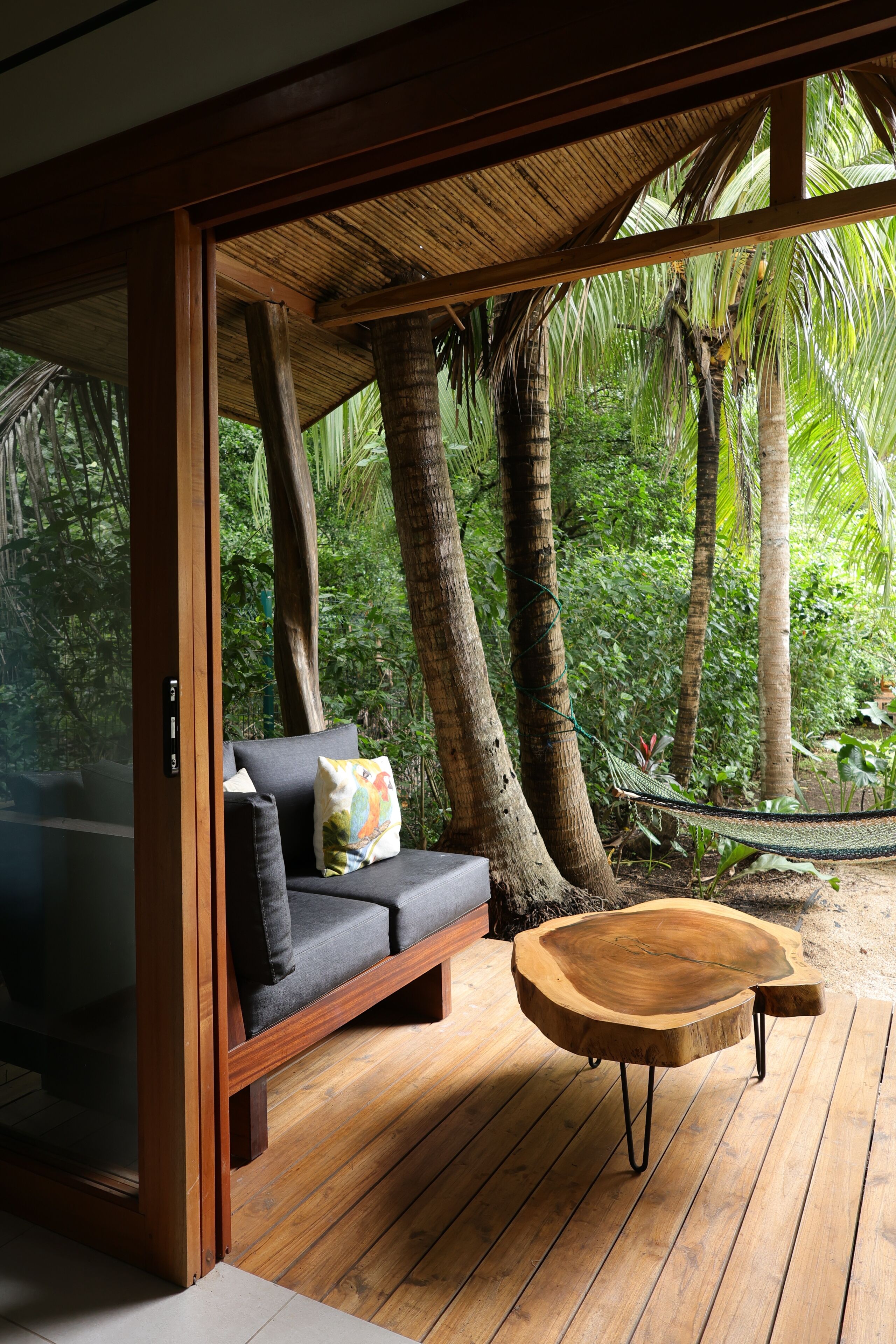 A wooden deck with a sofa and a round wooden table, surrounded by tropical vegetation.