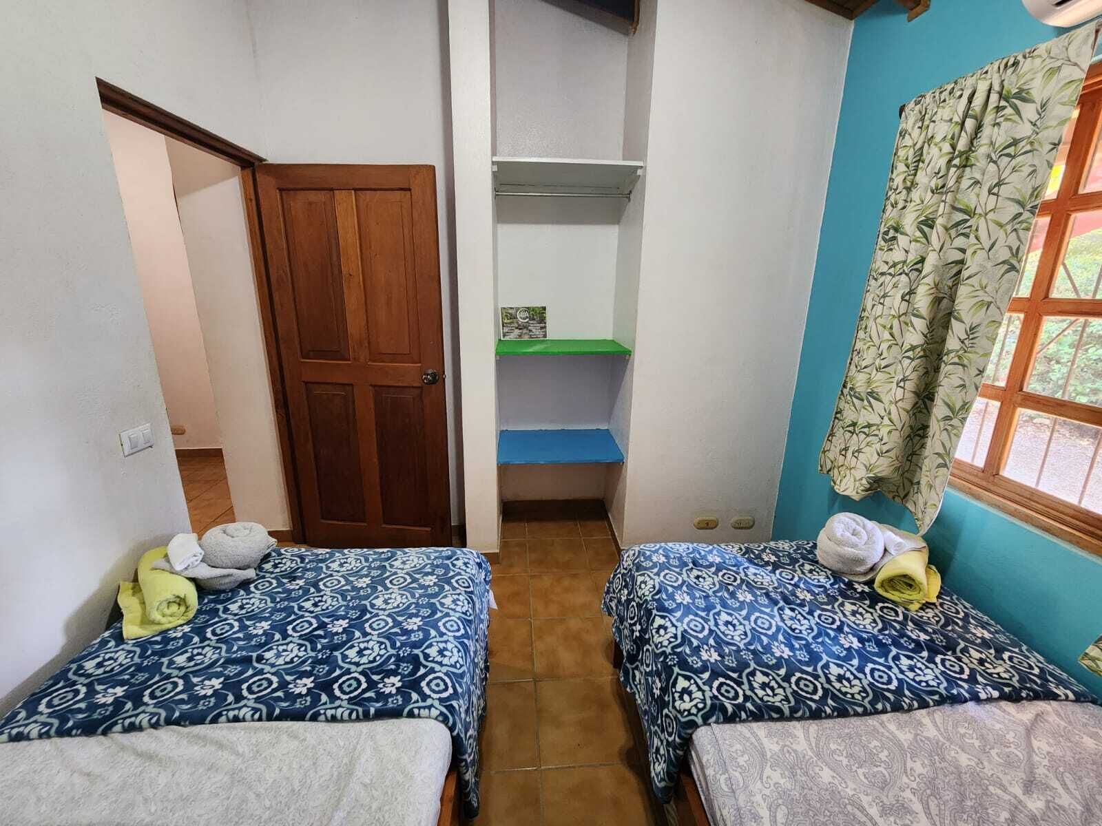 A small, narrow room with two beds, a wooden door, a shelf with a green top, and a window with patterned curtains.