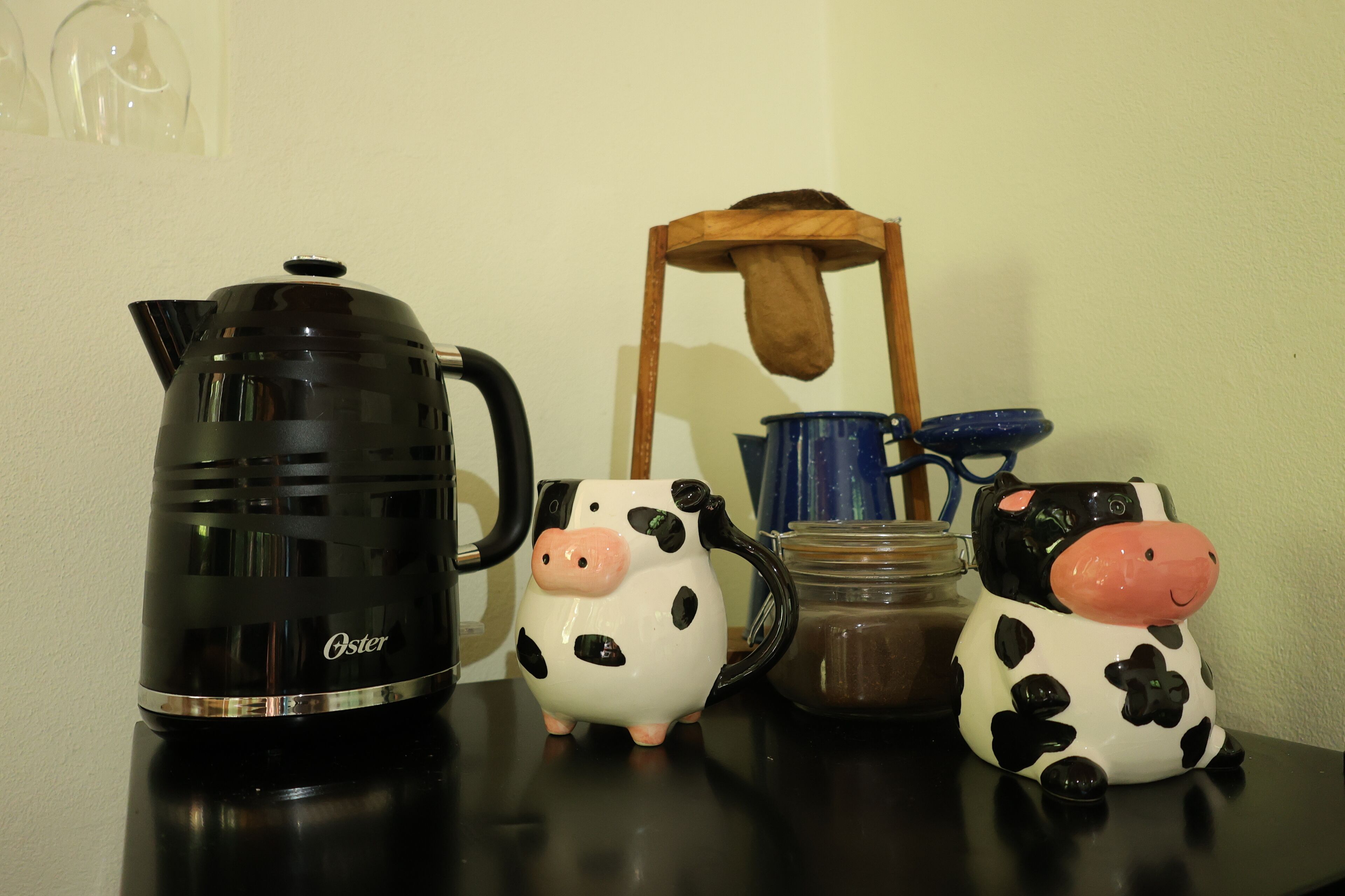 A black coffee maker, two ceramic pig-shaped mugs, a blue mug, and a wooden chair on a table.
