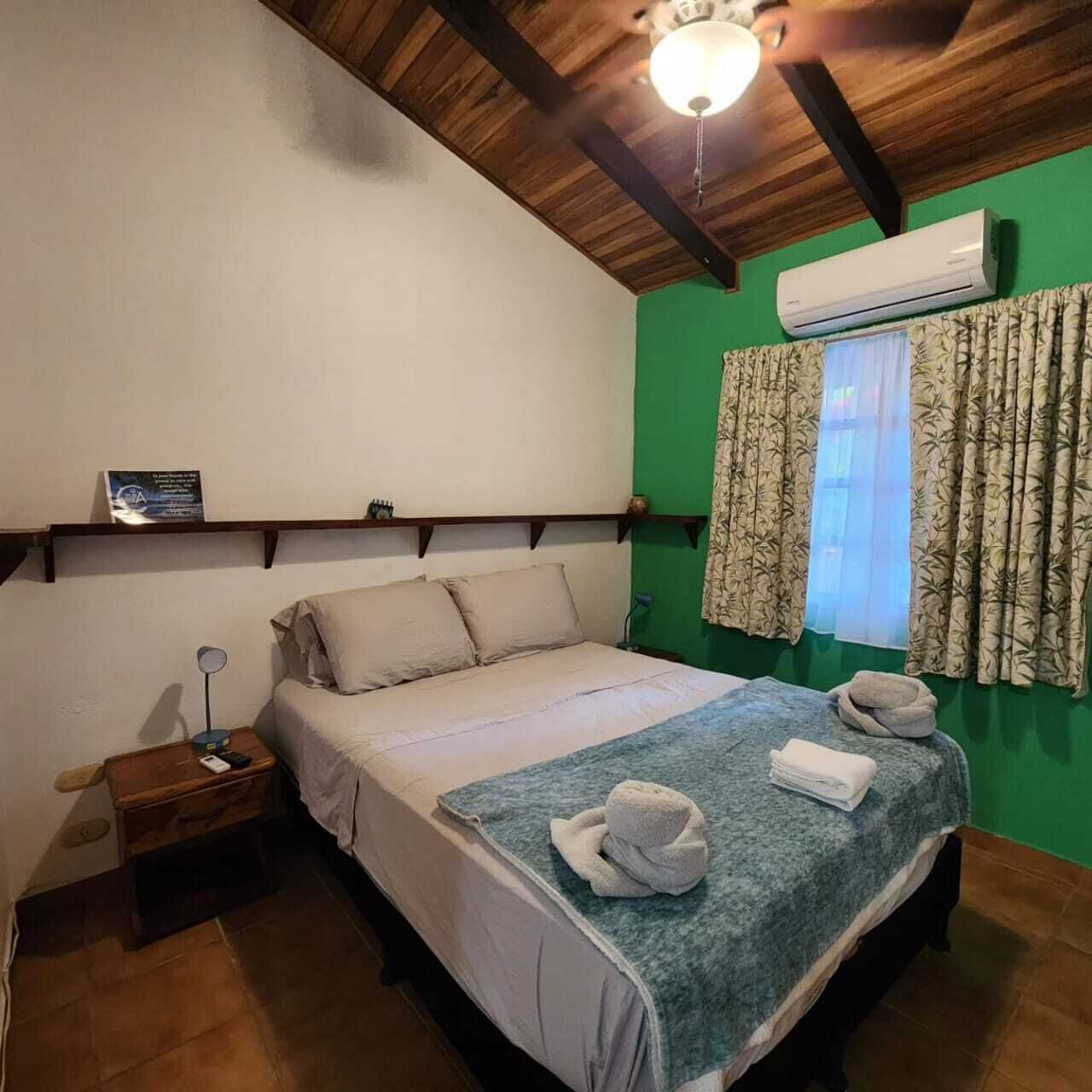 Un dormitorio con cama, mesita de noche, aire acondicionado, ventana con cortinas y ventilador de techo.