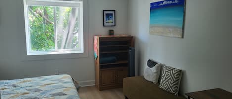 A room with a bed, a sofa, a small table, a bookshelf, a window with a view of trees, and a painting on the wall.