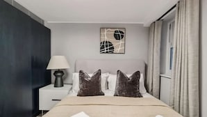 Habitación de hotel con cama, almohadas, mesita de noche, lámpara, una obra de arte en la pared y cortinas.